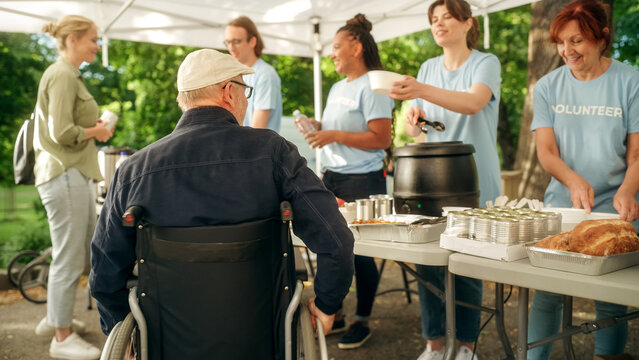 Positive And Smiling Middle Aged Man With Disabilities In A Wheelchair Receiving A Charity Meal From A Humanitarian Aid Food Bank. Volunteers Helping People In Need In Local Community.