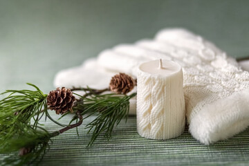 A white candle with a knitted pattern, white knitted gloves and a pine branch with cones. Card. Photo