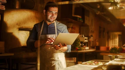 Fotobehang In Restaurant: Handsome Male Chef Using Laptop Computer. Authentic Pizza Place Cooking Delicious Organic Eco Food. Bi-racial male Entrepreneur Working on Online Order in Small Business Family Shop © Gorodenkoff