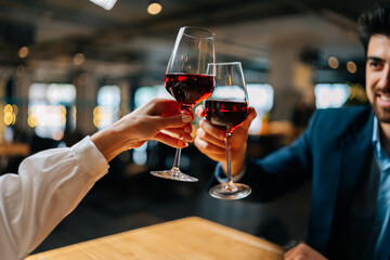 Close-up view from back of unrecognizable young woman clinking glasses of red wine with handsome smiling man sitting at table in restaurant at evening. Happy young couple enjoying nice romantic dinner
