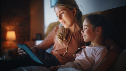 Mother Reading a Fairytale from a Tablet Computer to Her Lovely Daughter in Bed Before Going to Sleep. Young Mom Caring for Little Girl, Reading Online Stories at Home. Handheld Footage. © Gorodenkoff