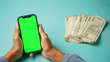 Top above flat lay view of a hand holding green screen mobile phone, Indian rupees currency over isolated background studio shot