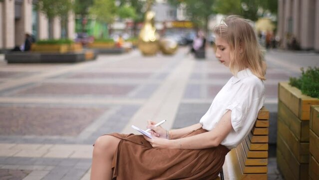 Side view of attractive focused female artist painting in sketchbook with pencil sitting on bench on modern city street at summer sunny day. Pretty young woman painting picture outdoors, slow motion.