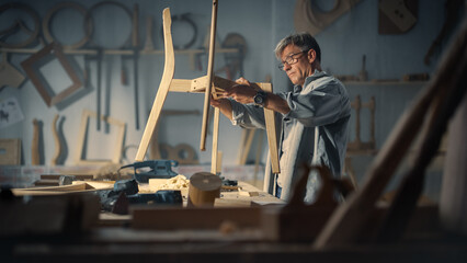 Adult Carpenter Putting on Glasses, Reading Blueprint and Starting to Assemble Parts of a Wooden Chair. Professional Furniture Designer Working in a Studio in Loft Space with Tools on the Walls.