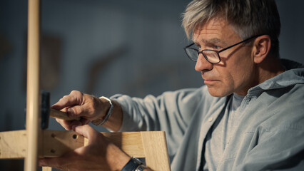 Adult Carpenter Putting on Glasses, Reading Blueprint and Starting to Assemble Parts of a Wooden Chair. Professional Furniture Designer Working in a Studio in Loft Space with Tools on the Walls.