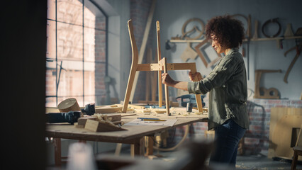 Young Stylish Female Carpenter Assembling a Wooden Chair. Professional Furniture Designer Working in a Studio in Loft Space with Tools on Walls Shot from an Angle Above.