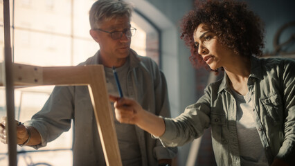 Obraz premium Close Up Portrait of Two Carpenters Working Together in a Carpentry Workshop. Multicultural Man and Female Colleagues Looking at a Blueprint on Paper and Discussing a New Chair Design.