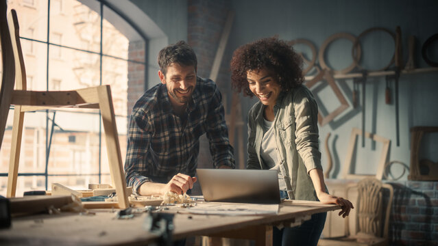 Happy Young Caucasian Man and Multiethnic Woman Using Laptop Computer in Designer Furniture Studio. Carpenters Have Fun, DIscussing and Talking About Upcoming Orders.