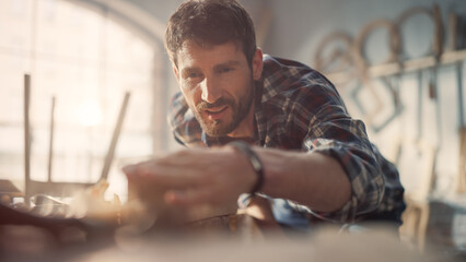Handsome Young Artisan Craftsman in Checkered Shirt Using Hand Plane to Shape a Wood Bar. Carpenter Working on a Project in a Loft Studio with Tools on Walls.