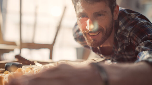 Talented Young Artisan Craftsman in Checkered Shirt Using Hand Plane to Shape a Wood Bar. Attractive Carpenter is Happy with the Results . Man Working on Project in a Loft Studio with Tools on Walls.