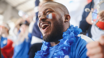Obraz premium Sport Stadium Event: Handsome Black Man with French Flag Painted on Face Cheering for His Team to Win. Crowd of Fans Shout Happily for the Scoring of the Goal. People Celebrate Championship Victory