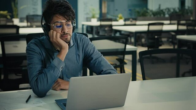 Bored tired arabian indian ethnic man businessman sit at office desk exhausted lazy sad male in headphones listen boring online webinar feel boredom lack of motivation dream about end of working day
