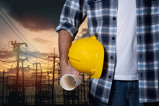 Engineer Man Construction Hand Holding Yellow Helmet Hardhat On Electric Pole At Power Plant Sunset Background. Worker Helmet Civil Construction Engineering. Close Up Engineer Hand Hold Safety Helmet.