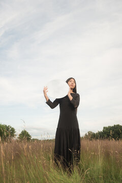 fine art portrait of happy woman in black dress standing in field holding round mirror reflecting cloudy sky