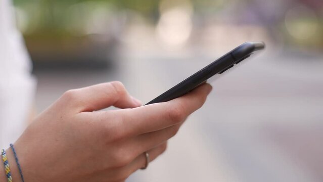 Close-up cropped shot of unrecognizable young woman messaging by mobile phone outdoors. Closeup of hands unknown female person touching smartphone screen on city street. Shooting in slow motion.
