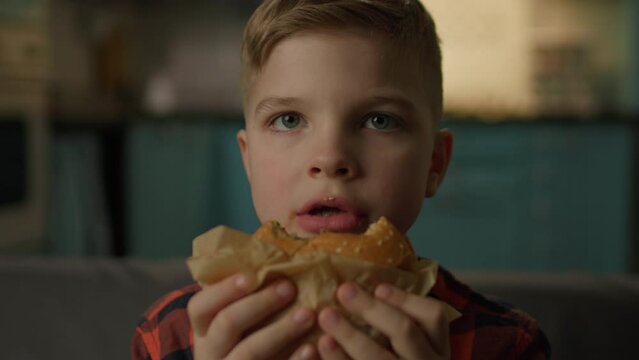 Hungry Kid Eating Burger Looking At Camera. Schoolboy chewing fast food burger sitting on sofa at home, close up.