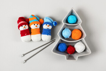 Three knitted snowmen in blue, orange and red hats with balls of thread and knitting needles on a gray background. Winter knitting creativity, the concept of New year and Christmas