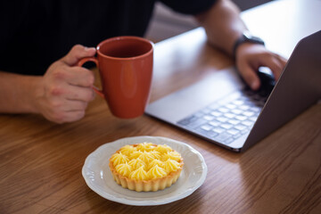 A white man wearing a black t-shirt working at home using a personal computer and holding a cup of coffee next to a delicious lemon pie during the day