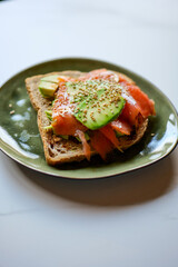 Fresh toast with avocado, salmon, vegan mayonaise, soya sauce and sesame seeds on green plate on marble kitchen top