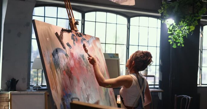 A woman artist in a spacious studio with panoramic windows makes strokes with a wide brush on large canvas on an easel. Talented female artist works on modern abstract oil painting, using paint brush.
