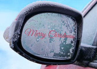 Red colour Merry Christmas handwriting or text on car side rear view mirror covered with frost against blurred snow and blue sky background, close up view.