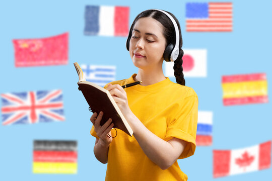 Language learning. A young Caucasian woman with headphones is writing something in a book. Blue background with defocused flags of different countries. Concept of education and university admission