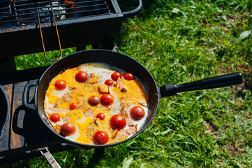 Fried scrambled eggs, bacon and cherry tomatoes in a cast iron frying pan, farm organic food cooked on open fire 