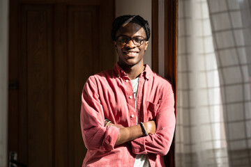 Smiling cheerful black millennial student guy standing in sunny room smiling at camera, enjoying dorm life. Happy young African man standing in cozy flat or apartment. Positive optimistic people