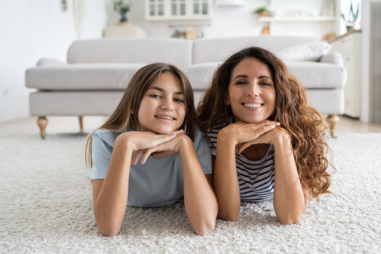 Charismatic cheerful woman and teen girl lying on floor with carpet posing for family photo. Positive contented school age daughter and millennial mom smiling looking at camera in living room