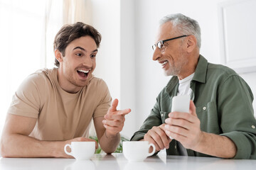 amazed guy pointing at mobile phone in hand of smiling dad while sitting near coffee cups in kitchen