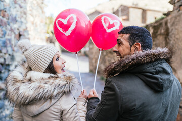 Young heterosexual couple looking at each other and laughing each other with a red balloon with white heart.