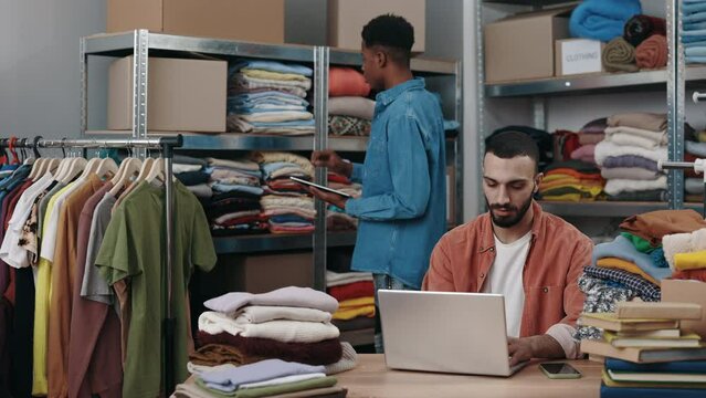 Male manager working with laptop in warehouse store while in clothing store. His multiracial colleague point at shelf counting with tablet at the background