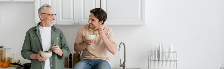 cheerful young man having breakfast and listening to positive dad in kitchen, banner