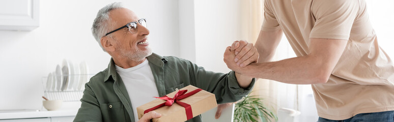smiling man holding gift box and shaking hands with son congratulating him on fathers day, banner