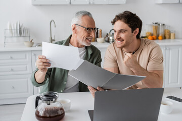 smiling mature man holding documents near excited son showing win gesture near laptop and coffee pot