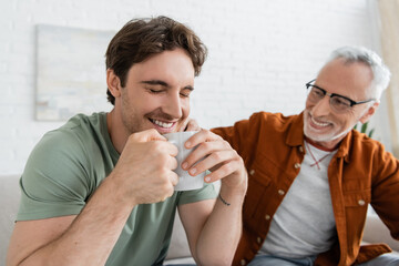 bearded and mature man talking to young son holding cup of tea while smiling with closed eyes