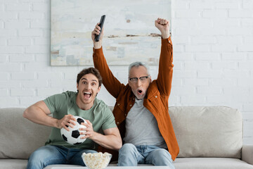 mature man showing win gesture and screaming near excited son holding soccer ball while watching...