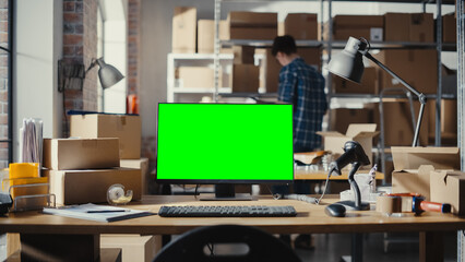 Desktop Computer Monitor Standing on a Table with a Green Screen Chromakey Mock Up Display. Small Business Warehouse with Worker Walking in the Background. Desk with Cardboard Boxes