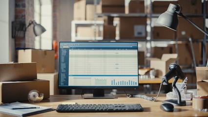 Desktop Computer Monitor Standing on a Table with Statistics and Data Display. Small Business Storage Room in the Background. Desk with Cardboard Boxes. Clients and Orders Infromation Management