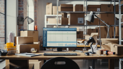 Desktop Computer Monitor Standing on a Table with a Display. Small Business Warehouse with Worker Walking in the Background. Desk with Cardboard Boxes