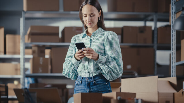 Portrait of a Beautiful Small Business Owner Using Smartphone at Work in a Warehouse. Female Connecting with Colleagues, Supporting Clients Over Internet. Sales Manager Working in Online Store.