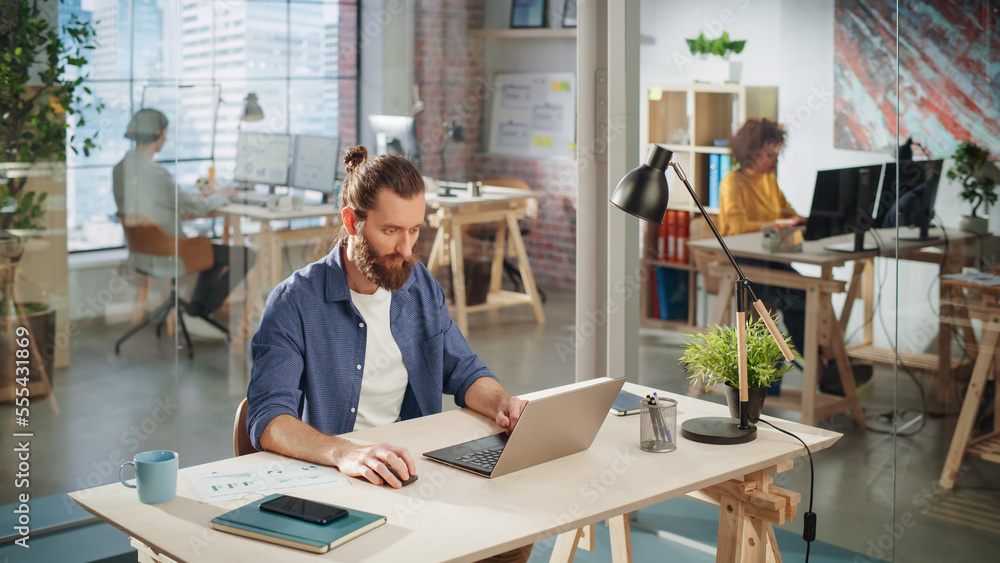 Aufkleber Handsome Long-Haired Bearded Manager Sitting at a Desk in Creative Office. Young Stylish Man Using Laptop Computer in Marketing Agency. Colleagues Working in the Background. High Angle View.