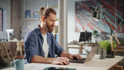 Handsome Long-Haired Bearded Manager Sitting at a Desk in Creative Office. Young Stylish Man Using Laptop Computer in Marketing Agency. Colleagues Working in the Background.