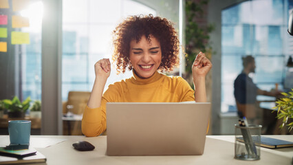 Portrait of a Beautiful Middle Eastern Manager Sitting at a Desk in Creative Office. Young Stylish Female with Curly Hair Celebrating her Achievement with Big Smile - Powered by Adobe