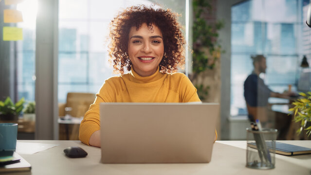 Portrait of a Beautiful Middle Eastern Manager Sitting at a Desk in Creative Office. Young Stylish Female with Curly Hair Looking at Camera with Big Smile. Colleagues Working in the Background.