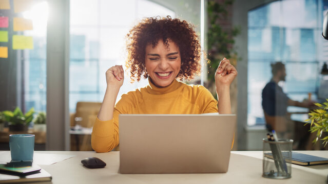 Portrait of a Beautiful Middle Eastern Manager Sitting at a Desk in Creative Office. Young Stylish Female with Curly Hair Celebrating her Achievement with Big Smile