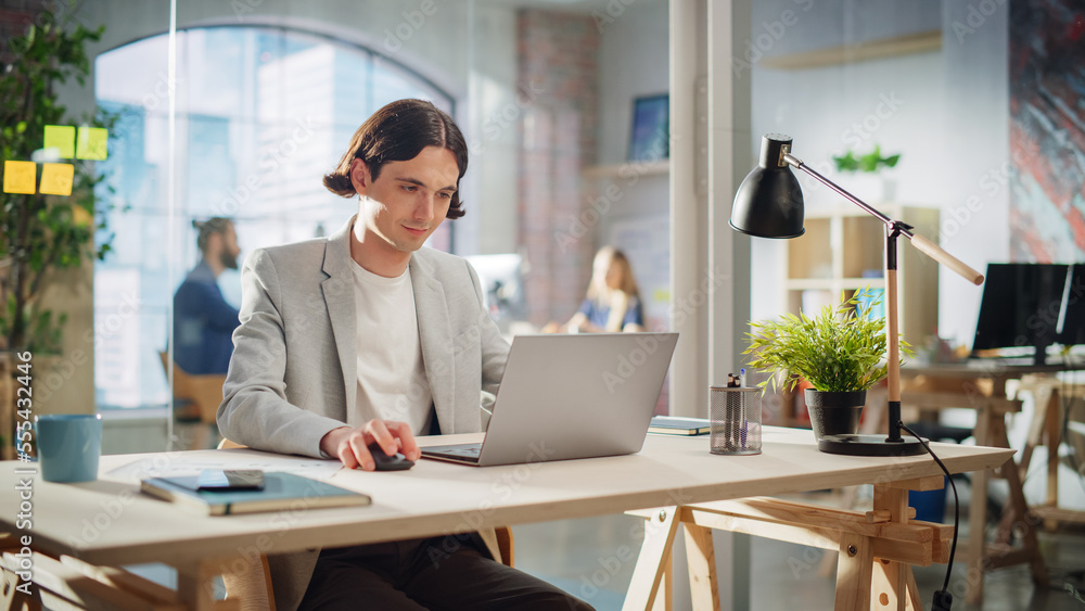 Poster happy long-haired manager sitting at a desk in creative office. young stylish man using laptop compu