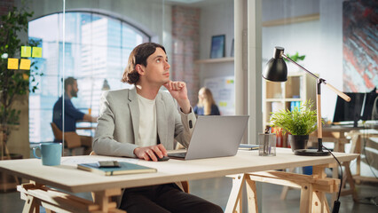 Handsome Long-Haired Employee Sitting at a Desk in Creative Agency. Stylish Man Using Laptop Computer in Marketing Company. Colleagues Working in the Background. Rack Focus Portrait.