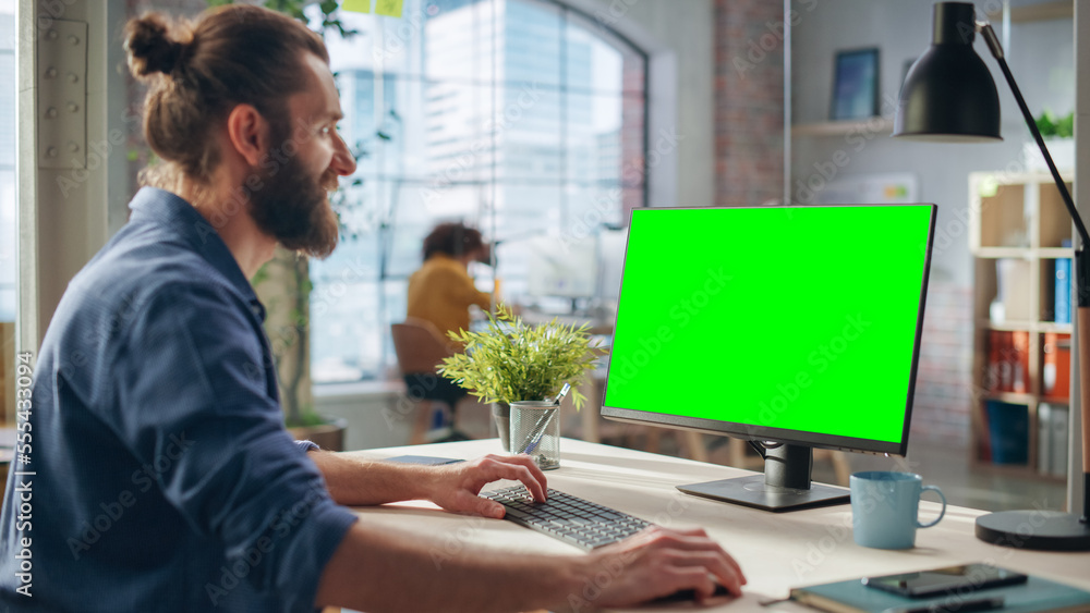 Poster Handsome Long-Haired Bearded Manager Working at a Desk in Creative Office, Using Desktop Computer with Green Screen Mock Up Display. Colleagues Working in the Background in Marketing Agency.