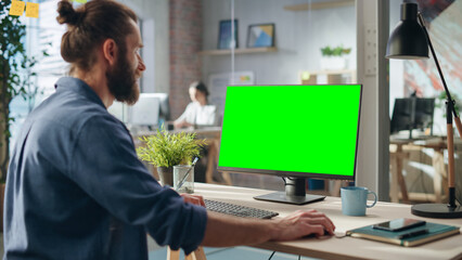 Handsome Long-Haired Bearded Manager Working at a Desk in Creative Office, Using Desktop Computer with Green Screen Mock Up Display. Colleagues Working in the Background in Marketing Agency.